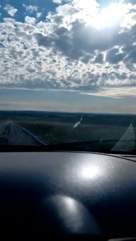 A first-person perspective shot from inside a vehicle looking out the windshield at a vast, flat landscape. The road ahead appears wet and stretches into the distance. Green fields and some bodies of water are visible on either side of the road. The sky is filled with dramatic, white, puffy clouds, with the bright sun shining through them. The dashboard of the car is visible in the foreground, reflecting the sunlight. The overall impression is of a drive through an open, possibly rural, environment under a dynamic sky.