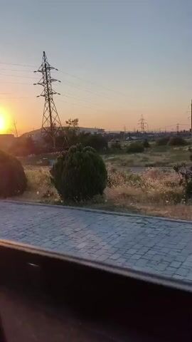 The video captures an outdoor scene during what appears to be sunset or early sunrise, given the warm, golden light illuminating the sky and landscape. The perspective suggests the video is filmed from a moving vehicle, possibly a car or bus, looking out the side window.

Dominating the left side of the frame is a tall, metallic electricity pylon with multiple crossbars and wires stretching across the scene. The pylon is silhouetted against the sky. In the foreground, a paved walkway made of grey interlocking bricks is visible, bordered by a concrete curb.

The surrounding environment is semi-arid with patches of dry grass and scattered shrubs. Several bushes, some rounded and well-maintained, and others more natural and wild, are interspersed throughout the landscape. A few trees, including a tall cypress-like tree on the right, are also present.

In the middle ground, beyond the vegetation, there are signs of development. A few low-rise buildings or structures can be seen, along with more electricity pylons and wires in the distance, suggesting an industrial or suburban area. A dark-colored car is parked in the distance on the left side of the frame. The sky is clear with a gradient of colors from a pale orange near the horizon to a light blue at the top. The overall impression is one of a landscape transitioning from day to night or vice versa, with man-made infrastructure coexisting with nature.