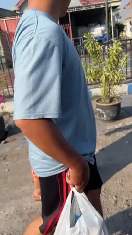 The video shows a person walking while holding a white plastic bag. The person is wearing a light blue t-shirt and black shorts with red stripes. They are walking on a dirt or gravel path with a plant in a pot to their right. In the background, there are buildings and a street. The lighting suggests it is daytime.