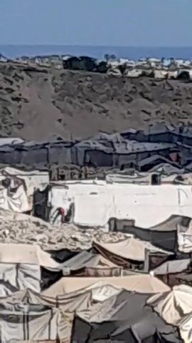 The video shows an aerial view of a densely populated refugee camp. Tents and makeshift structures are visible, made of canvas and other scrap materials. In the foreground, a horse-drawn cart carrying two people moves along a dirt path. In the middle ground, several individuals are seen walking between the tents. In the background, a barren, hilly landscape with more structures can be observed. The overall scene depicts a community living in challenging conditions, likely due to displacement.