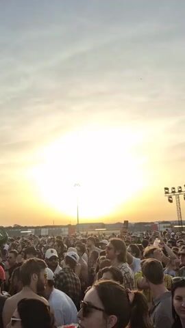 Here's a description of the video:

The video shows a large crowd of people at an outdoor music festival during a sunset. The sun is low in the sky, creating a warm glow over the scene. People are densely packed together, facing towards the stage. Some individuals are wearing hats and sunglasses.

In the background, there's a large passenger airplane. A stage is set up in front of the airplane. There are tall light fixtures, part of the concert setup, visible throughout the crowd. The sky is mostly clear.