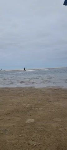 A person kite surfing on a body of water with a sandy beach in the foreground. The sky is overcast with a light grey cloud cover. The water appears calm with small ripples. In the distance, a sandbar or narrow strip of land separates the body of water from what appears to be the ocean. There are other people visible on the sandbar, some standing and some appearing to be participating in water activities. The overall mood is serene, suggesting a peaceful day at the beach for water sports. The lighting is diffused, typical of an overcast day. No visible text is present in the image. The location context of Naaldwijk, Netherlands, places this scene along the Dutch coastline.