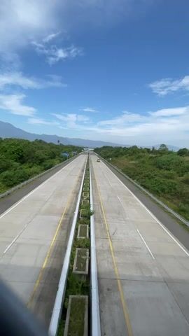 A vertical, eye-level, outdoor, daytime shot captures a wide, multi-lane highway stretching into the distance under a bright blue sky with scattered white clouds. The highway has two sets of lanes, separated by a concrete median with planters filled with greenery. Yellow lines demarcate the lanes, and the road surface appears to be concrete. To the left and right of the highway, rolling green hills covered in dense trees form the landscape. In the distance, a faint mountain range is visible. A few vehicles are present on the highway: a yellow car in the distance on the right lane, a black car in the left lane, a silver car in the left lane, a dark car in the distance on the left lane, and a yellow truck on the right lane. The camera is positioned from an elevated viewpoint, possibly a bridge or overpass, as suggested by a dark railing or obstruction in the very bottom foreground. The overall atmosphere is serene and open, suggesting a clear and pleasant day for travel.