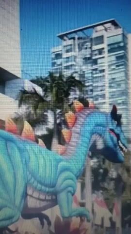 The video shows a bright blue dinosaur figure with orange spikes along its back. The dinosaur is in the foreground, with a tall, modern building and palm trees in the background. The sky is clear and blue. In the second half of the video, a person is seen from behind, looking out at the scene. A white carousel horse is also visible in the foreground.
