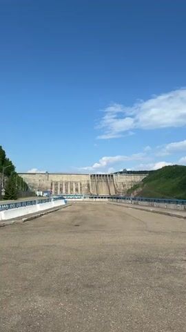 A person walks across a paved area with a blue railing on their left. In the distance, a large concrete dam is visible, with lush green hills behind it and a clear blue sky with scattered clouds above. The camera pans to show more of the dam and the surrounding landscape. Another shot shows a wide river flowing between densely forested hills.