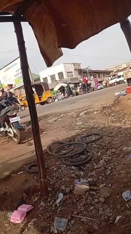The video shows a busy street scene, likely in an urban area of Africa. From a partially obscured vantage point under some kind of shelter, one can see a variety of activity taking place. In the foreground, there's a parked motorcycle, a pile of discarded tires, and some litter. A man wearing a blue shirt and dark pants walks away from the camera, crossing the street. Further down the street, there are several auto rickshaws, motorcycles, and pedestrians. A billboard advertising A.A. Rano Rice Mill is visible in the background. The scene is slightly dusty, with a mix of paved and unpaved surfaces.