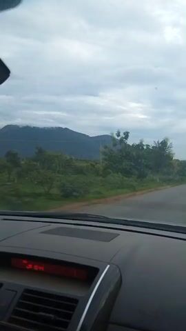 A passenger's view from inside a car dashboard, looking out the windshield on a cloudy day. The road curves to the right in the foreground, leading towards a hazy mountain range in the background. Lush green vegetation covers the landscape, with scattered trees and some agricultural fields. A telephone pole is visible on the left side of the road. A motorcycle with two riders is seen in the distance on the right side of the road, approaching the car. The car's dashboard has a red digital display with numbers.