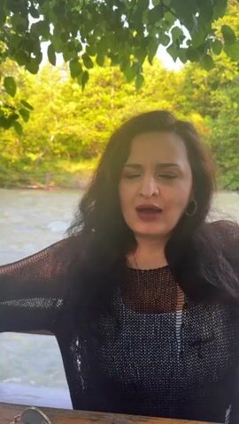 The video shows a woman with long, dark hair sitting outdoors near a body of water. She is wearing a dark, sheer sweater over a patterned top. The woman is speaking and gesturing with her hands, her facial expressions conveying strong emotion. The background features green foliage and sunlight filtering through the leaves, creating a warm and natural ambiance. The focus is on the woman, with the environment serving as a backdrop.