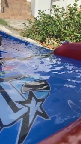 A close-up shot focuses on a blue and red inflatable pool filled with water. A person's hand and foot enter the water from the left side of the frame. The camera angle shifts slightly as the person's body becomes more visible in the pool, with their legs submerged. In the background, a green leafy plant grows near a white structure and some stairs. The sunlight is bright, indicating an outdoor setting. The person is playing in the pool.