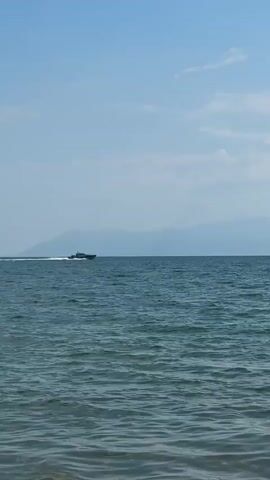 A speedboat moves across a body of water. The boat is dark colored and appears to be moving at a fast pace, leaving a white wake behind it. The water is choppy with visible waves. In the distance, a hazy mountain range is visible against a clear blue sky with scattered white clouds. The camera is positioned low, close to the water's surface, with the foreground showing ripples of water.
