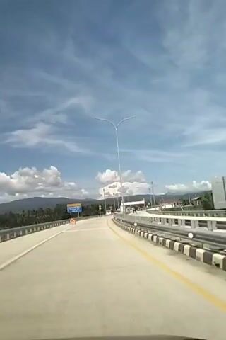 The video shows a first-person view of driving on a highway towards a toll plaza. The sky is blue with some white clouds. There are mountains and trees in the background. The road is paved with white lines and a yellow line on the right side. There are guardrails on the right side of the road. There are streetlights and signs visible. The toll plaza has multiple booths and some vehicles are visible. The video is from 00:00 to 00:05.