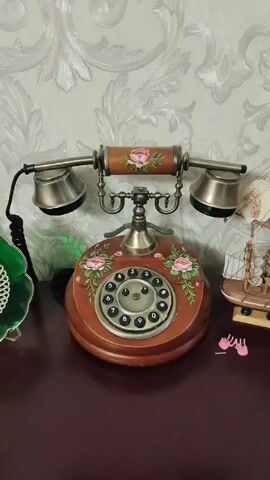 A vintage rotary phone, adorned with hand-painted pink roses and green leaves, sits on a dark wooden surface. The phone has a circular dial with black numbers and symbols, a metal headset, and a wooden body. A person's hand, with painted pink fingernails, is visible, interacting with the phone by touching the dial and the base. To the left of the phone, a green decorative object with a white lacy pattern is partially visible. To the right, a small model sailboat is present, along with a pair of tiny pink high-heeled shoes. The background features a light-colored wallpaper with a raised, ornate floral pattern. The scene appears to be indoors, likely in a room or on a shelf, with artificial lighting. The activity is focused on the interaction with the telephone. There are no discernible time of day or weather cues, and no visible text. The overall impression is one of a decorative display or a playful interaction with an antique item.