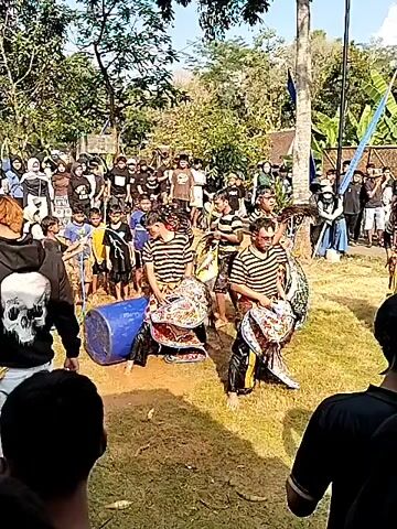 A group of performers in black and white striped shirts and colorful patterned skirts are dancing in an open grassy area, some are playing drums. A crowd of onlookers stands behind them, watching the performance. The environment appears to be outdoors, with trees and a bamboo fence visible in the background. The sun is shining, casting shadows on the ground. Some of the performers are wearing masks.