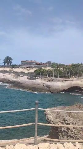 The video shows a scenic view of a rocky coastline with the ocean and palm trees. In the foreground, there is a railing made of rope and metal posts, separating a stone pathway from the cliff edge. The sea is a vibrant blue, with waves gently lapping against the rocks. In the background, there is a large building, possibly a hotel, situated on a hilltop surrounded by palm trees. The sky is blue with some scattered clouds. The video pans slightly to the right, revealing more of the coastline and the open sea. There are a few small boats visible in the distance on the water.