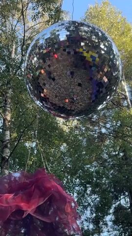A large, mirrored disco ball hangs suspended from a wire outdoors, surrounded by trees with green foliage. Below the disco ball, a large, dark pink, translucent fabric decoration is visible. The sky is a clear blue, indicating daytime. The disco ball is reflecting the surrounding environment. The video appears to be a static shot with slight natural movement from wind.
