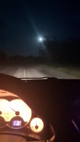 A nighttime scene from inside a moving vehicle shows the view through the windshield. A road stretches out ahead, illuminated by the vehicle's headlights. The sky is dark, with a bright full moon visible above. Trees line the sides of the road. A utility pole is visible on the right. The vehicle's dashboard is visible at the bottom of the frame, showing the speedometer and other gauges, which are dimly lit. The vehicle appears to be traveling at approximately 50 kilometers per hour. Another vehicle’s lights are seen in the distance.