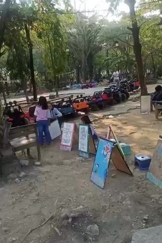 A group of children and adults are in an outdoor park-like setting. In the background, a line of small, colorful toy electric cars is parked. In the foreground, several children are interacting with colorful A-frame display boards, some of which appear to be for drawing or art activities. Some children are sitting or standing near the display boards, and one child is being attended to by an adult. The ground is sandy and dirt. Trees and foliage are visible throughout the scene, suggesting a natural environment.