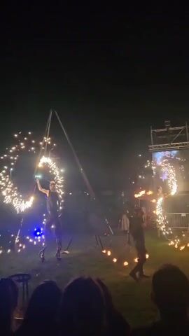 A medium shot captures a nighttime fire show with three performers in a grassy field. On the left, a performer in a dark top and light pants operates a large, sparkling firework contraption in a spiral shape, illuminating the dark night sky. In the center, a performer in a white garment stands amidst several small fires on the ground. On the right, another performer in a dark top and pants manipulates a chain of burning torches, creating large flames. In the background, a large stage with a screen displays moving images. A dark audience is visible in the foreground.