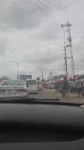 The video shows a view from inside a car driving down a street. The sky is overcast and a tall communications tower is visible on the right. There are several cars and small buses ahead of the vehicle, appearing to be in slow-moving traffic. On the right side of the road, people are walking along the side of the street, some carrying umbrellas. There is a mix of structures visible including what appears to be shops or stalls along the side of the road. The interior of the car including the dashboard and side pillar are visible. The scene appears to be in a busy area, possibly a market or commercial district in a developing country.