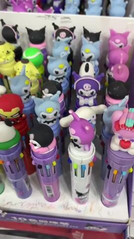 The video pans across a display of stationery, primarily featuring novelty pens and pencil sharpeners. The first section shows a collection of multi-colored pens topped with cartoon characters like My Melody and Kuromi, available in various pastel shades. In the next shot, Hello Kitty themed pencil sharpeners are displayed in a pink box, alongside smaller pastel-colored sharpeners. Further along, more multi-colored pens are visible in a tray, along with small packages of colorful clips. The video concludes with a view of a shelf stocked with pencils in a metal holder and more stationery items in packaging. The overall environment appears to be a retail store or a stationery shop.