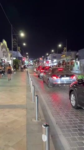 A nighttime view of a busy street with cars in traffic and pedestrians on the sidewalk. The street is lined with buildings displaying lights and signs. The road is paved with brick-like stones, and metal bollards separate the sidewalk from the road. Several cars are stopped, their brake lights illuminated. Pedestrians are walking along the sidewalk, some are looking at their phones. The sky is dark and clear.