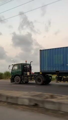 A semi-trailer truck drives down a road. The truck is green, and it is pulling a large blue shipping container. The sky is cloudy. The road appears to be a highway.