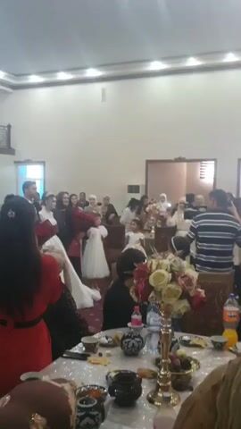 A wedding reception is taking place in a large, indoor venue with high ceilings and ornate lighting. The bride and groom are central figures, dressed in traditional wedding attire. The bride wears a white gown, and the groom a suit. They are surrounded by guests, many of whom are dressed formally. Several people are holding bouquets of flowers, including the bride and groom. In the foreground, a table is set with food, drinks, and decorative items. Some guests are seated at tables, while others stand and observe the proceedings. A few children are present, dressed in formal clothing. In the background, there is a balcony with people standing and watching. The overall atmosphere appears festive.