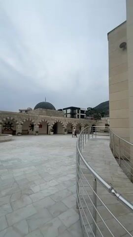 The video shows an outdoor courtyard with a paved floor made of white marble tiles. In the background, there is a building with arched walkways and a dome, likely a mosque. Two people are walking in the courtyard. A curved ramp with a metal railing is visible on the right side of the frame. The sky is overcast.