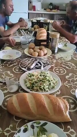 The video shows a table laden with food. There are four people visible, sitting around the table, eating. The food includes bread, potatoes, fish, and salad. The room appears to be a kitchen or dining area. The lighting is natural, coming from windows. The overall atmosphere is one of a shared meal.