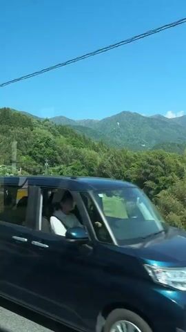 The video shows a scenic view from a moving vehicle.  A gray guardrail runs parallel to the road on which the vehicle is traveling. Beyond the guardrail, there's a lush green landscape featuring trees, vegetation, and a valley. In the distance, there are mountains visible under a clear blue sky. Some clouds are seen hovering above the mountain range.