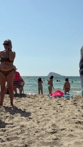 A woman in a brown bikini stands on a sandy beach, holding a phone to her ear and looking to the left. Behind her, several people are visible in the water and on the shore. In the distance, a hilly island is visible in the ocean under a clear blue sky. A man in a blue shirt and grey shorts stands on the right side of the frame, facing away from the camera.
