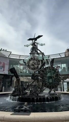 A metal sculpture of a bird is in the center of a fountain, with water spraying from the base. The sculpture is made up of gears, chains, and other mechanical parts, with a large clock face in the center. The sculpture is surrounded by a circular walkway with shops in the background. The sky is cloudy.