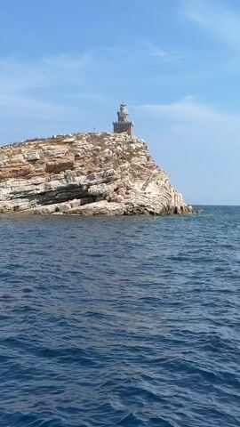 This media file captures a serene coastal scene featuring a lighthouse perched atop a rocky island. The lighthouse, constructed from stone, stands tall and proud against a backdrop of a clear blue sky with scattered white clouds. The island itself is a rugged formation of layered rock, with sparse vegetation clinging to its surface. A set of stairs winds its way up the side of the island, leading towards the lighthouse, hinting at a challenging but rewarding ascent.

The foreground is dominated by the deep blue, rippling waters of the sea, suggesting the perspective is from a boat. The sunlight glints off the water's surface, indicating it is likely daytime. The overall atmosphere is one of tranquility and natural beauty, with the lighthouse serving as a solitary sentinel against the vastness of the ocean. There are no people visible in the media file. The location is identified as Portoferraio, Italy. The time of day appears to be afternoon, given the bright sunlight and clear sky.