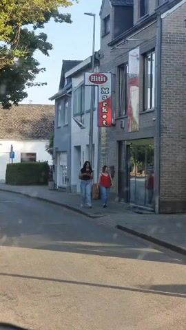The video shows a street scene in Heusden-Zolder, Belgium. Two young women are walking on the sidewalk in front of a brick building. The woman in the foreground is wearing a dark brown top and light blue jeans, and she is looking at her phone. The woman behind her is wearing a red tank top and light blue jeans.

The building appears to be a storefront, with large glass doors and windows. A red and white sign is visible above the entrance, with the word "Hilit" in white letters on a red background, and below it, "Market" in red letters on a white background. The time of day appears to be late afternoon or early evening, as the sun is casting long shadows on the street and sidewalk. The weather seems pleasant, with clear skies and sunlight.

The scene is calm and ordinary, with no indication of any particular event or urgent activity. The women are walking at a casual pace, suggesting they are going about their daily business. The background shows other buildings and trees, indicating a typical urban or suburban environment. There are no cars visible on the street, and the sidewalk is clear except for the two pedestrians. The overall impression is one of a quiet, sunny day in a European town.