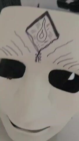 The video begins with a close-up of a mask, which appears to be white with a black drawing on it. Next, a black handgun is shown resting on a surface next to a sword. The scene then shifts to a wider view of a living room. A plush tiger toy is lying on the floor. In the background, a person is standing in front of a mirror, holding a phone and taking a selfie. The person is wearing black shorts and dark shoes. The room appears to be well-lit, with a sofa and a coffee table visible.