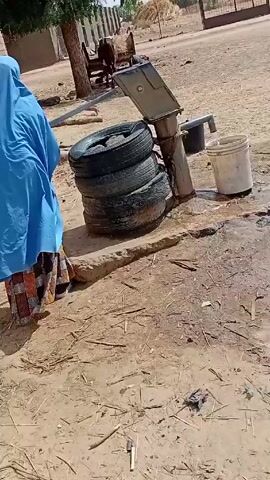 The video shows a hand-operated water pump with tires stacked around its base filling a white bucket with water. A woman in a blue hijab and patterned skirt walks past the pump, followed by a young child in a blue striped outfit. The ground is dry and dusty with scattered straw. In the background, there is livestock, a fence, and other buildings. The scene appears to be in a rural or agricultural setting.