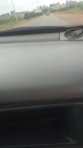 The video provides an interior view from a vehicle's dashboard looking out through the windshield. The road ahead is visible, with a single car in the distance. The environment appears to be rural or suburban, with sparse vegetation and a cloudy sky. The dashboard itself is gray, and a small black compartment is visible below it. A circular emblem with a star-like design is affixed to the dashboard near the top right. The video appears to be a continuous shot with no discernible events or interactions occurring.