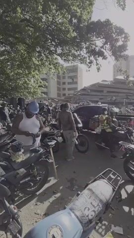 The video shows a street scene with several people on motorcycles. In the background, a large building has collapsed, with debris piled high. The sky is bright and sunny, with trees providing some shade. Some people are wearing masks. The overall impression is one of a disaster zone with people navigating the area on their motorcycles.