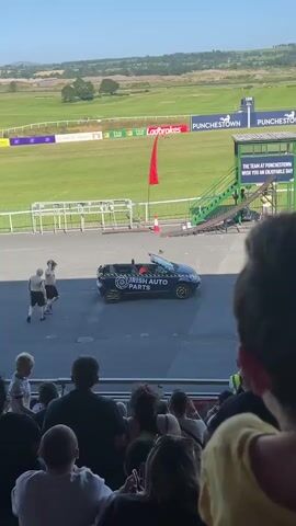 The video captures a sunny outdoor event at what appears to be a racecourse. In the foreground, a crowd of spectators is seated on bleachers, looking towards the action. In the midground, a convertible car with "IRISH AUTO PARTS" branding drives in a semi-circular motion. The car is dark colored with yellow accents on its wheels and has two people visible inside. A ramp structure is set up near the car. A person on a bicycle is seen to the left of the car. In the background, a vast green field stretches out with various advertisements and flags visible, suggesting a large-scale event. The overall atmosphere is festive and lively.