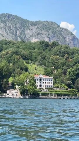 A wide shot captures a grand white villa with an orange-tiled roof situated on a lush green hillside overlooking a shimmering blue lake. The villa is flanked by manicured gardens and trees, with a steep, rocky mountain rising majestically behind it. The sky is a clear blue with a few wispy clouds. The water in the foreground shows ripples, suggesting gentle movement. The scene appears to be set in a serene and picturesque location, likely a sunny day given the bright lighting and clear sky. The overall impression is one of opulent beauty and tranquility, characteristic of a luxury lakeside resort or private estate. The time of day appears to be midday or early afternoon. There is no visible text in the media file.