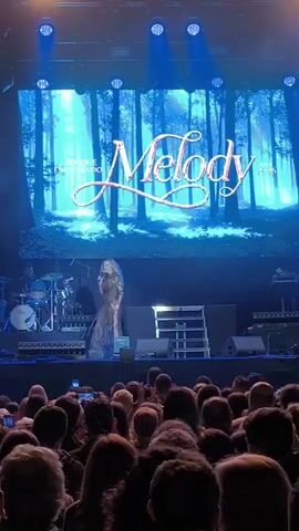 A female vocalist sings onstage while the audience records her with their mobile phones. The stage is bathed in blue light, and a screen behind the singer displays the word "Melody" in cursive against a background of trees. The artist wears a long gown and has blonde hair. A drummer is visible behind her to the left, and other band members are present. The audience, composed of adults, faces the stage, holding their phones up to capture the performance. There are stage lights and a small set of steps on the stage. The overall impression is that of a live music event.