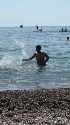 A young person is playing in the water near a beach. The person is splashing and appears to be having fun. In the background, there are other people on paddleboards and a boat. The water is clear and blue, and the sky is bright and sunny. The beach is covered in small stones.