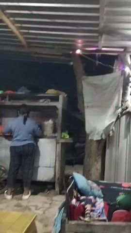A woman in a blue shirt and patterned pants stands facing away from the camera, working at a food stall. Beside her, a man in a blue shirt and dark pants is cooking at a wok over a gas burner. The stall is covered by a corrugated metal roof supported by wooden beams. A yellow tarp covers a table in the foreground. The scene is dimly lit and appears to be outdoors at night.