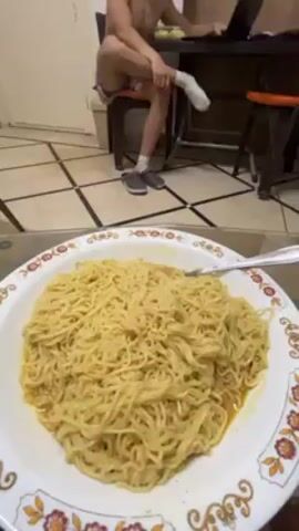 A video shows an interior scene. A plate of yellow noodles sits in the foreground. The camera pans to reveal a young man seated at a desk in the background. He is shirtless and wearing patterned shorts, one white sock, and gray sandals. He is sitting with one leg crossed over the other and is working on a laptop. There is a partially visible room divider behind him. The floor is tiled with a light color, and black tape divides the tiles into squares.