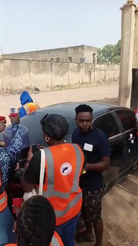 The video shows a group of people gathered around a dark-colored car. Most of them are wearing orange safety vests. One man in a green shirt is pouring a purple liquid from a large water bottle into small red cups. Several other people are holding cups out to receive the liquid. The car's trunk appears to be being used as a makeshift table.  The group is in an outdoor area with a paved surface and a tall concrete wall in the background. Some individuals in the crowd are wearing face masks. The weather appears to be sunny.