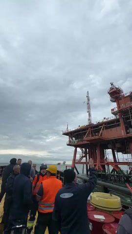 A group of people are gathered on the deck of a boat, observing a large, red offshore oil or gas platform under an overcast sky. Some individuals are wearing high-visibility vests and hard hats, suggesting they are workers or visitors to the industrial site. Red barrels are visible on the boat's deck in the foreground.

The platform is a multi-level structure with various equipment, including a tall mast-like feature and cranes. In the distance, another smaller platform can be seen. A helicopter flies into view from the left side of the frame, moving across the platform, indicating potential transport or surveillance activity.

One man in the foreground, wearing a dark jacket with "AAM STORNOWAY" visible on the back, points towards the platform. The scene depicts an industrial offshore environment, likely near Marina di Ravenna, Italy, given the provided context. The time of day is daytime, with grey, cloudy weather. Text "SC...W-C" is partially visible on the platform.