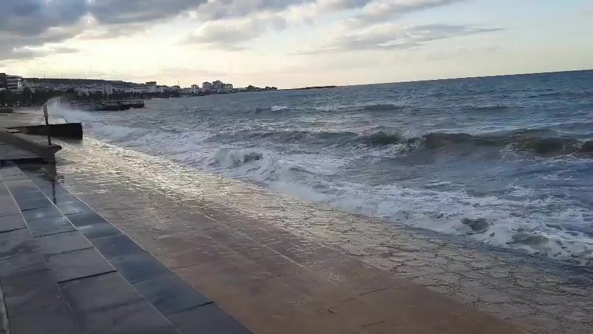 The video shows a turbulent sea with large waves crashing against a concrete promenade. The sky is overcast with clouds. In the distance, a city skyline is visible. The waves are powerful, with white foam and spray. The concrete steps and promenade are wet, indicating the waves are reaching the edge. There are no people or animals visible in the video.