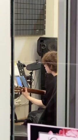 A young person with curly brown hair, wearing a black shirt, is seated in what appears to be a music studio or practice room. They are playing an acoustic guitar, holding its neck with their left hand and strumming with their right. Two smartphones are placed on a music stand in front of them, seemingly displaying sheet music or tabs that the person is reading.

Behind the person, parts of an electronic drum set and a large speaker or amplifier are visible. Acoustic foam panels are mounted on the wall. The scene is viewed through a glass panel, as indicated by a horizontal line of reflection across the frame. The room is equipped with various musical instruments and sound-treatment materials, suggesting a dedicated space for music. No specific time of day or emotions are discernable. The location context provided is Селекционер-2 Снт, Russia.