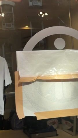 The scene is a close-up, indoor shot of a glass door or window. A white sticker or decal is affixed to the glass, depicting a stylized design consisting of an arc over a circle. The sticker is partially covered with pieces of translucent white paper or plastic, secured by tan-colored tape. Behind the glass, the reflection of an exterior building is visible. Miscellaneous items are clustered near the bottom of the frame, including luggage, bags, and discarded items. Lighting fixtures are visible in the reflection.