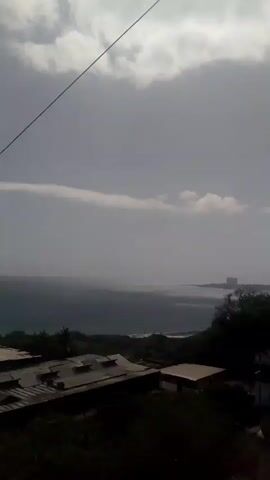 The video captures an overcast view of the ocean from a slightly elevated position overlooking some buildings. The sky is filled with gray clouds, with a thin band of lighter clouds stretching horizontally across the horizon. The sea appears dark and choppy, with waves breaking near the shore. In the foreground, there are several low-rise buildings with flat roofs, some of which appear to be in disrepair. The surrounding area is covered in lush green foliage, suggesting a tropical or subtropical environment. The overall mood of the video is somber and perhaps a bit desolate due to the weather and the condition of the buildings.
